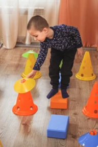 A boy arranges orange traffic cones and blue foam blocks on a wooden floor as part of an indoor obstacle course 