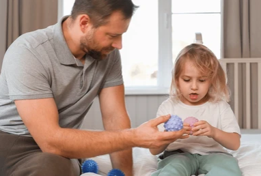 Father helping young girl play with textured balls on a bed in a bright room 