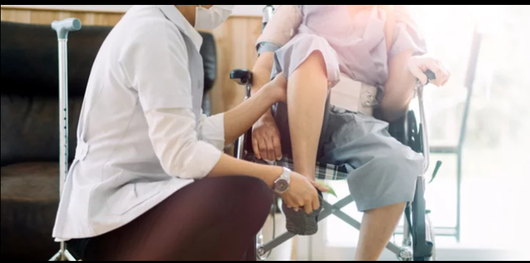 Caretaker in white coat assisting a child in a wheelchair, adjusting the child's shoe while seated indoors 