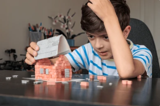 Boy concentrating as he assembles a small brick toy house on a table, with loose bricks nearby 