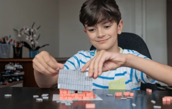 A smiling boy in a striped shirt stacks pink bricks and a grey roof on a small model house at a desk 