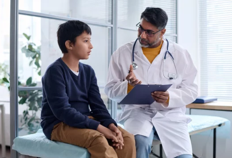 Doctor wearing a white coat with a stethoscope speaks with a young boy seated on an examination bench in a clinic, holding a clipboard in hand 