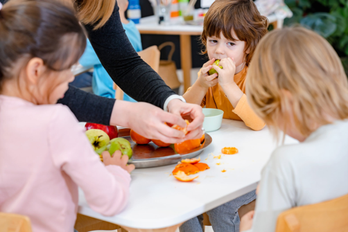 Children peel and snack on oranges at a white table, guided by an adult in a bright classroom kitchen setting 