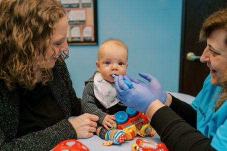 Infant sits on a table during a pediatric checkup as a clinician in blue gloves examines the baby's mouth with parents watching kindly nearby 